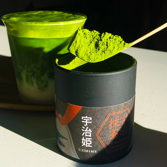 Matcha green tea powder in a black container with a spoonful of powder above it, and a cup of matcha in the background. Ujihime by Chikiriya Tea House